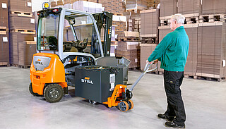 A man uses a hand pallet truck to pull a large battery sideways out of a forklift in a warehouse.