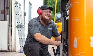 Ein Servicetechniker mit Gehörschutz und Schutzbrille kniet vor einem STILL Stapler um ihn zu reparieren