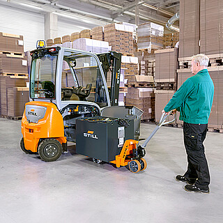 Ein Mann zieht mit einem Handhubwagen eine große STILL Blei-Säure-Batterie seitlich aus einem Gabelstapler in einer Lagerhalle.