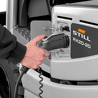 Nahaufnahme einer Hand, die einen Ladestecker in die seitliche Ladebuchse eines silbernen Gabelstaplers (Modell STILL RX20-20) steckt.