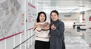 Zwei Frauen stehen in einem KION Büro und beide formen jweils ein Herz mit einer ihrer Hände