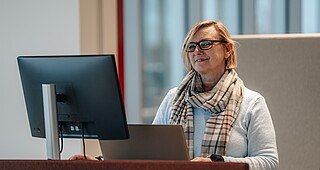 Eine Person steht in eienm Büro vor ihrem LAptop an den ein zweiter Bildschirm angeschlossen ist.
