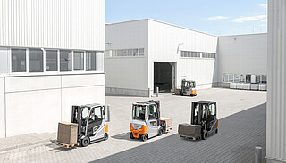 Four forklifts transport loaded wooden pallets on a paved industrial site in front of white industrial buildings.