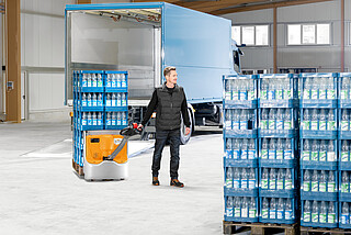 Mann zieht mit elektrischem Hubwagen Paletten mit Wasserflaschen in einer Lagerhalle vor einem blauen Lieferwagen
